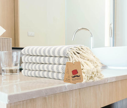 Four grey Arpa cotton hand towels stacked beside a bathroom sink with cream tassels and a Bazaar Anatolia tag.