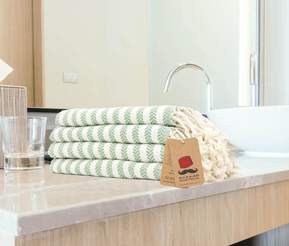 Four folded green Arpa cotton hand towels beside a bathroom basin, showing woven stripes and cream tassels.