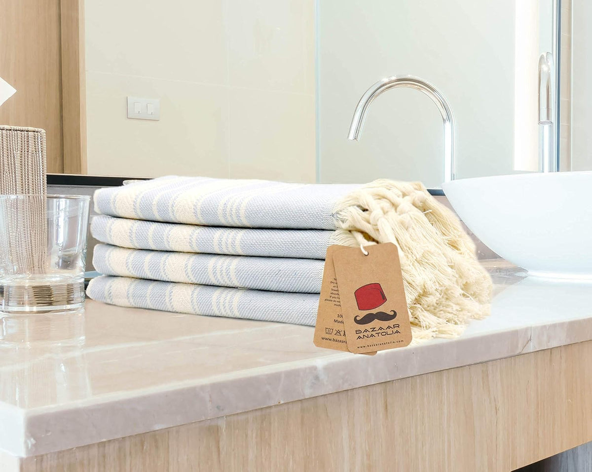 Four folded light blue Stripe cotton hand towels stacked beside a bathroom basin with cream tassels.