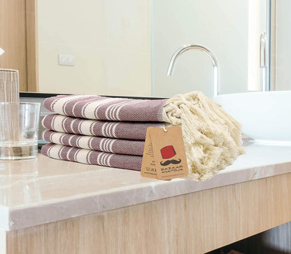 Four folded maroon Stripe Turkish hand towels stacked beside a bathroom basin with cream tasseled ends.