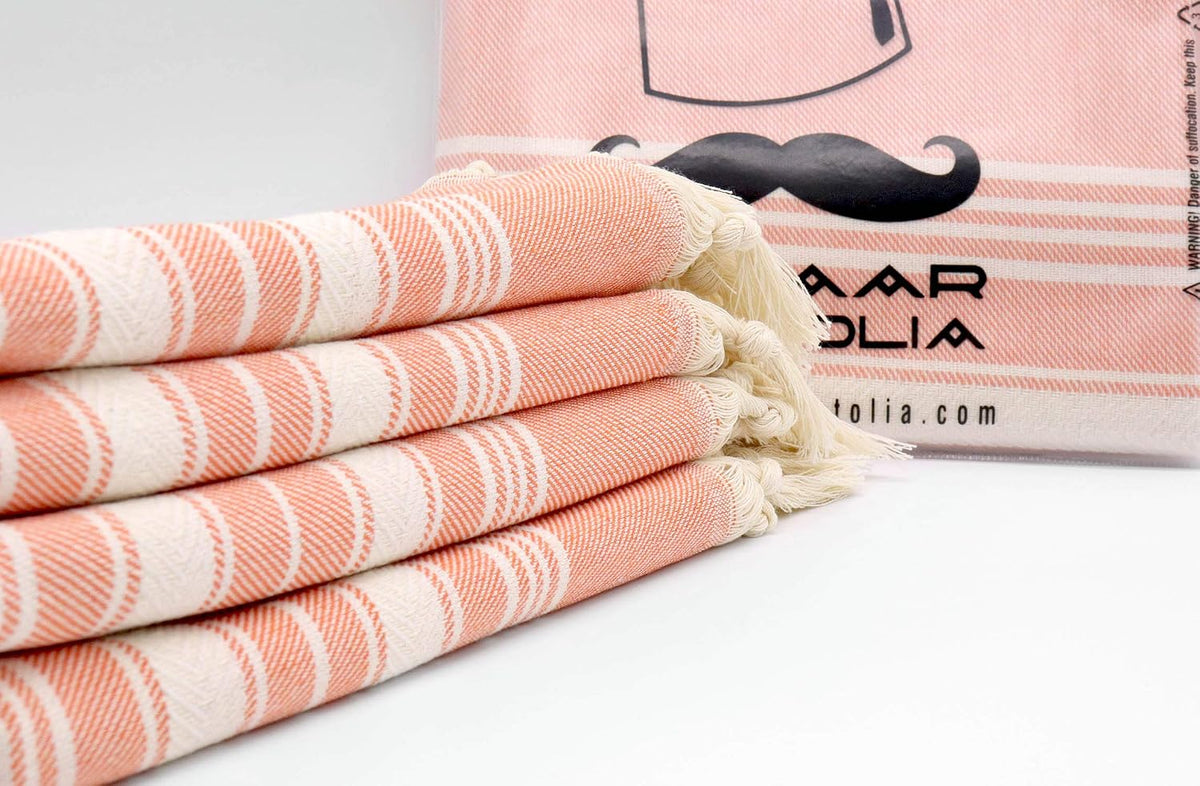 Close-up of four folded orange Stripe cotton hand towels with cream tassels beside a Bazaar Anatolia display.