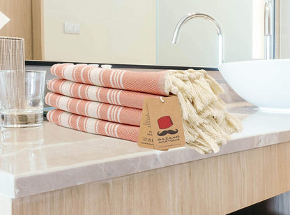 Four orange Stripe cotton hand towels with cream tassels stacked on a bathroom counter.