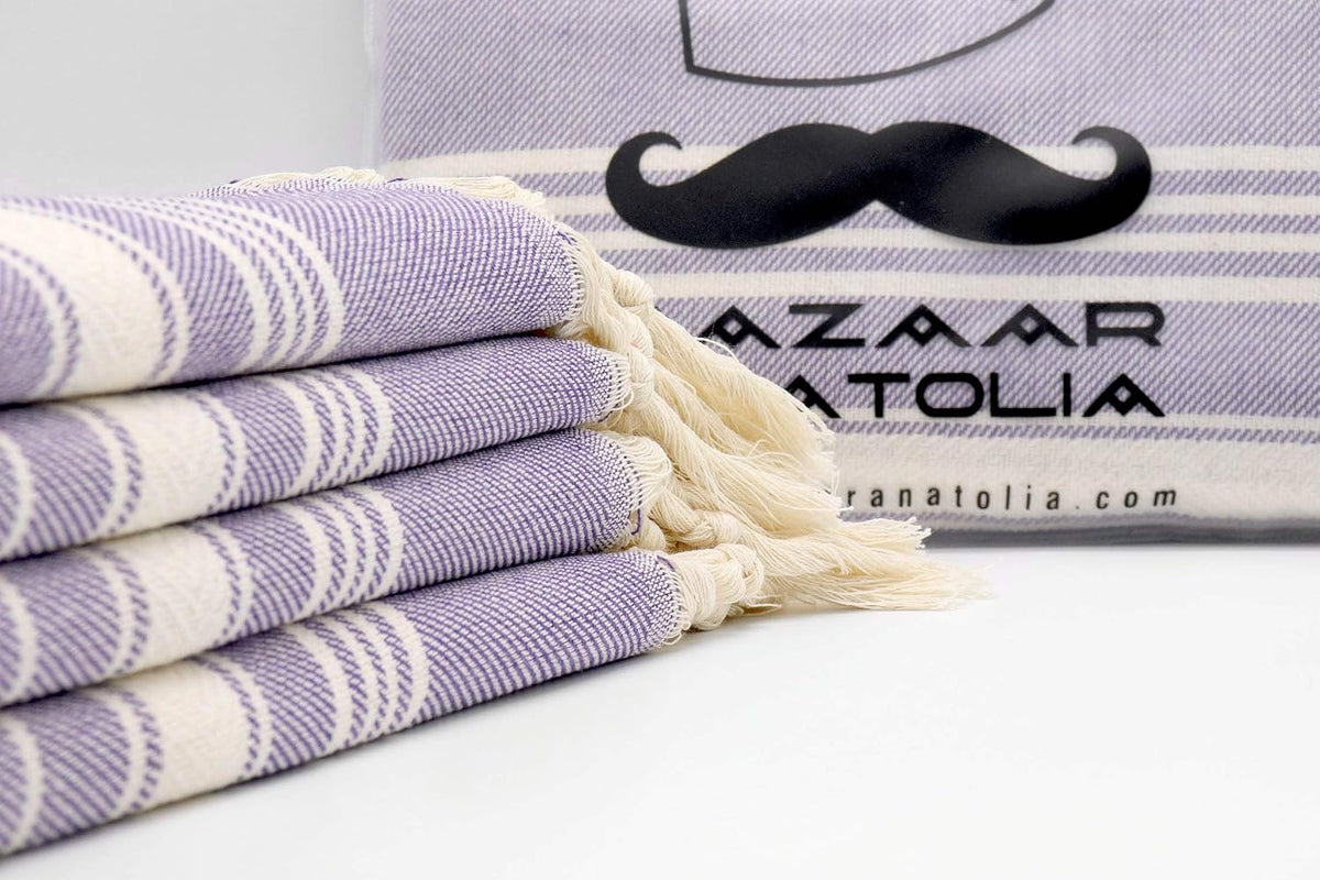 Four folded purple Stripe Turkish hand towels with cream tassels shown close up beside a Bazaar Anatolia display.