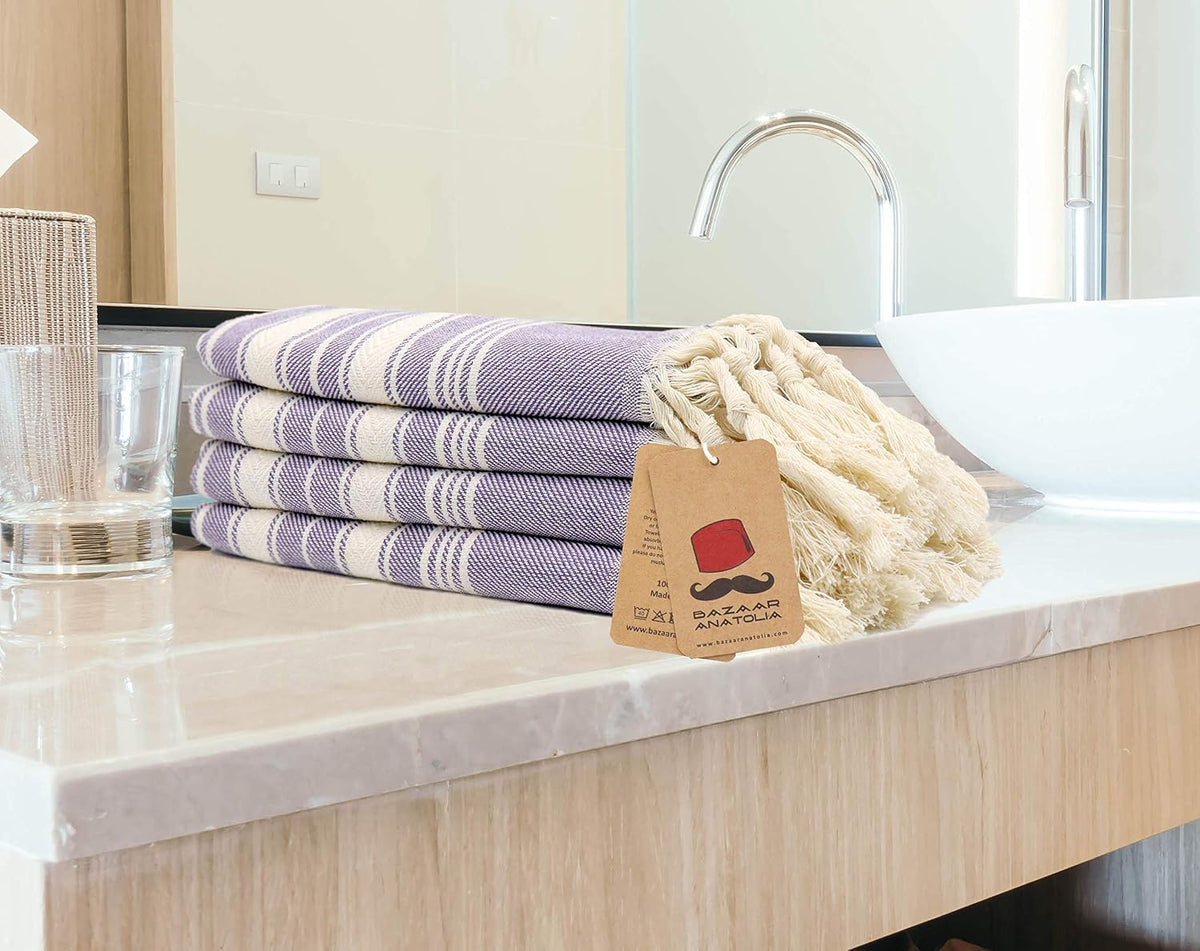 Four purple Stripe Turkish hand towels stacked beside a bathroom basin with cream tasseled ends.