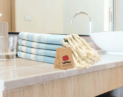 Four turquoise Stripe Turkish cotton hand towels stacked beside a bathroom sink with cream tassels.