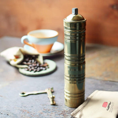 Handmade brass Turkish coffee grinder with detachable crank handle, shown with coffee beans, a cup of coffee, and a branded Bazaar Anatolia storage pouch for a traditional coffee brewing experience.